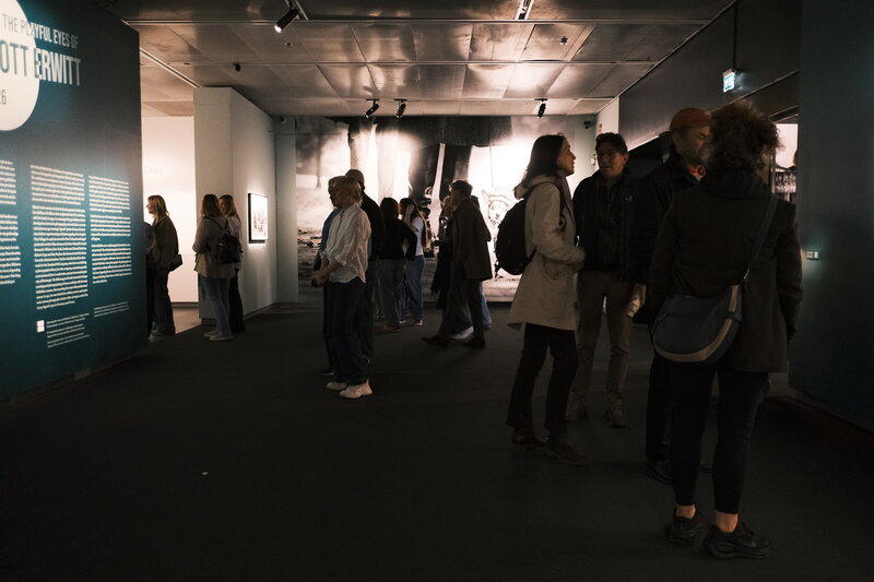 Visitors browsing a photography exhibition, large black-and-white prints on dark walls, text panels visible on the left