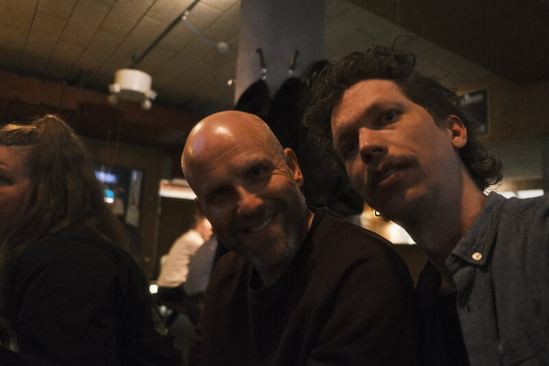 Two men leaning in close together for the camera at a bar — one bald and smiling broadly, one with curly hair and a moustache